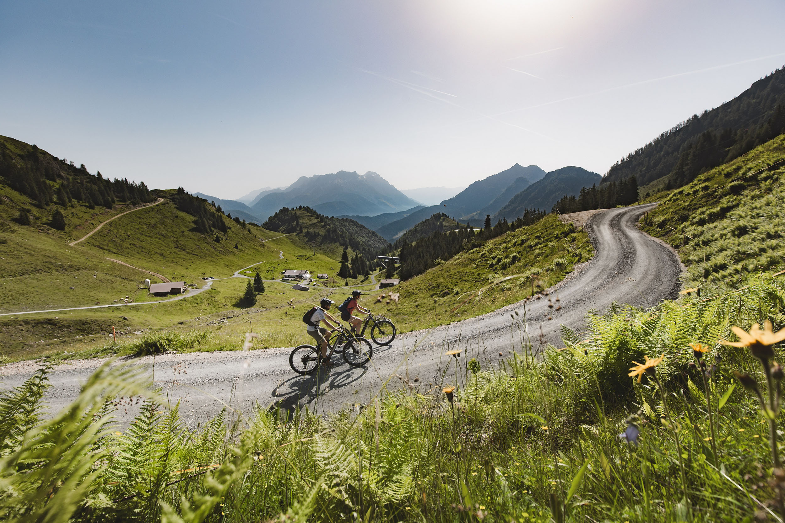 Zwei Radfahrer fahren auf einem kurvigen Schotterweg durch eine sonnige Berglandschaft im Pillerseetal.
