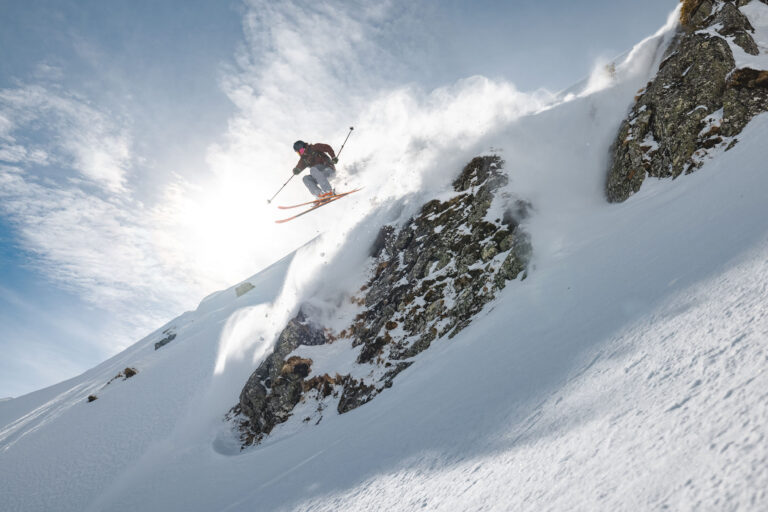 Ein Freerider springt dynamisch über einen felsigen Hang und wirbelt dabei Schnee auf.