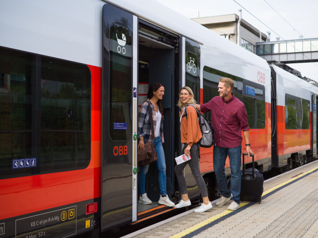 Drei junge Menschen steigen entspannt an einem Bahnhof aus einem modernen ÖBB-Zug. Das Bild symbolisiert klimafreundliche und nachhaltige Mobilität mit der Bahn in Österreich.