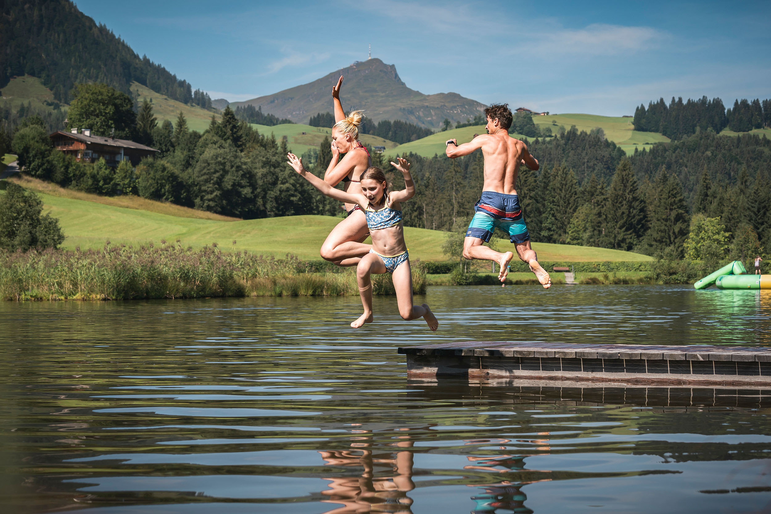 Eine Familie springt an einem Sommertag fröhlich von einem Steg in den Naturbadesee Lauchsee in Fieberbrunn.