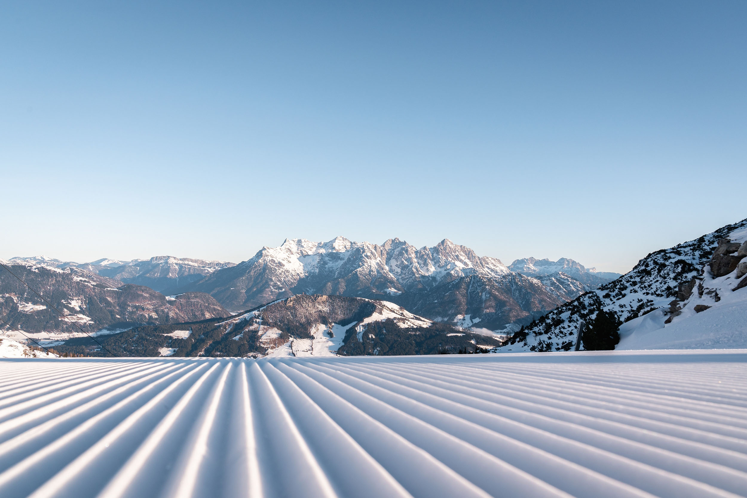 Perfekt präparierte Skipiste mit Blick auf die verschneiten Loferer Steinberge unter klarem, blauem Himmel im Skigebiet Saalbach Fieberbrunn.