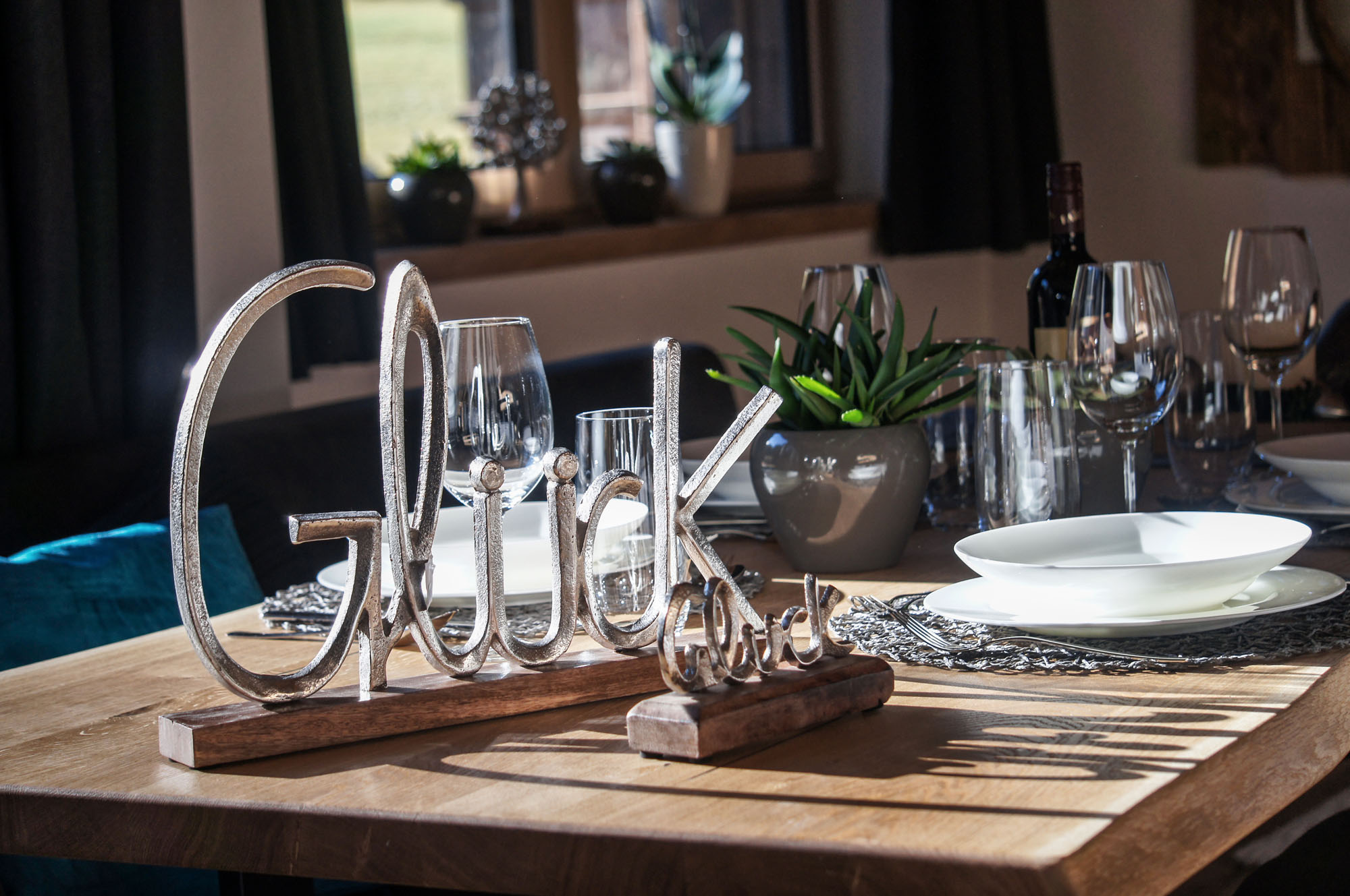 Detail eines gedeckten Holztischs mit Deko-Schriftzug „Glück“, weißen Tellern, Gläsern und Sukkulenten im Chalet-Stil