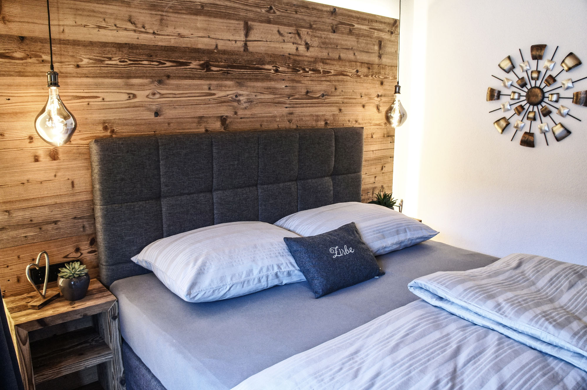 Stilvoll eingerichtetes Schlafzimmer mit einem Boxspringbett in Grau, flankiert von zwei modernen Hängelampen mit Vintage-Glühbirnen. Eine rustikale Holzverkleidung bildet das Kopfteil, davor ziert ein Zierkissen mit der Aufschrift „Zirbe“ das Bett. Nachttische aus naturbelassenem Holz mit dekorativen Elementen und eine wandmontierte metallene Stern-Dekoration ergänzen das harmonische Design.