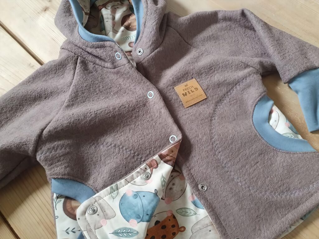 Babyjacke von "Rosas Handarbeiten" aus Fieberbrunn, handgenäht. Die Jacke ist aus weichem, braunem Fleece gefertigt, hat hellblaue Bündchen und Druckknöpfe. Die Kapuze sowie das Innere sind mit einem bunten Stoff mit Tiermotiven gefüttert. Auf der Vorderseite befindet sich ein Lederpatch