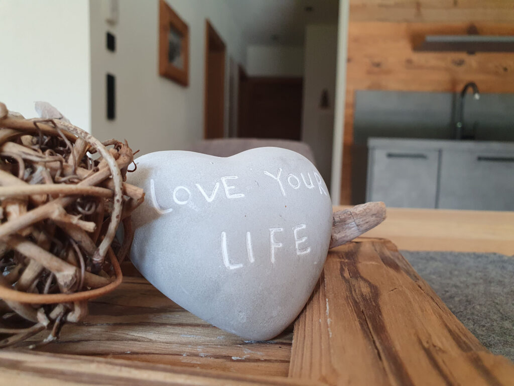 Nahaufnahme Herzstein „Love Your Life“ auf Holztablett vor Chaletküche