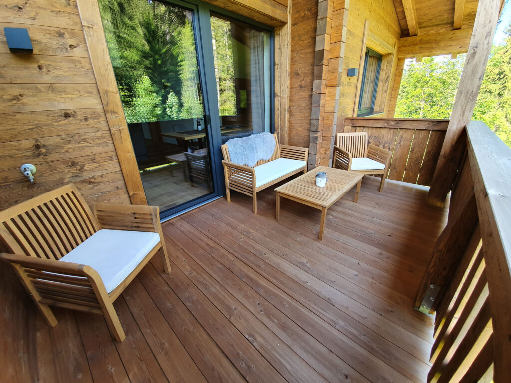 Geräumiger Holzbalkon mit Liegestühlen, Couchtisch, Alpenwaldkulisse und ruhiger Sommerstimmung