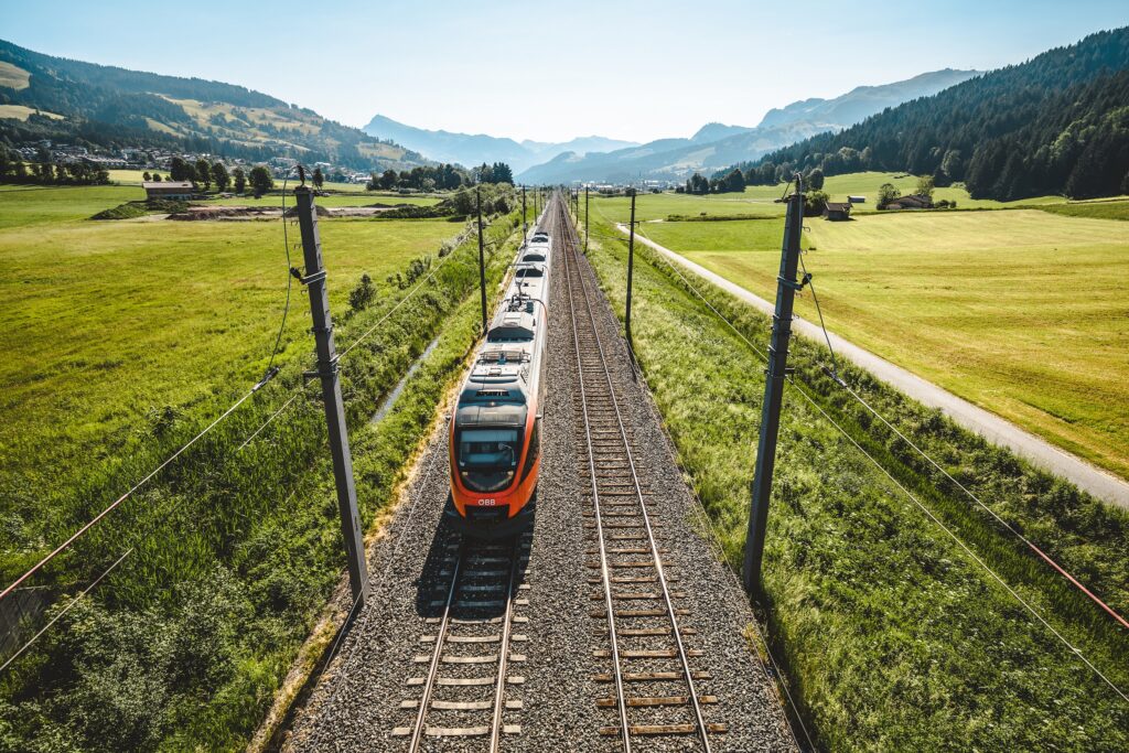 Ein ÖBB-Zug fährt durch eine grüne, hügelige Landschaft in den Alpen. Das Bild steht für nachhaltige, umweltfreundliche Mobilität und klimafreundliches Reisen in Österreich