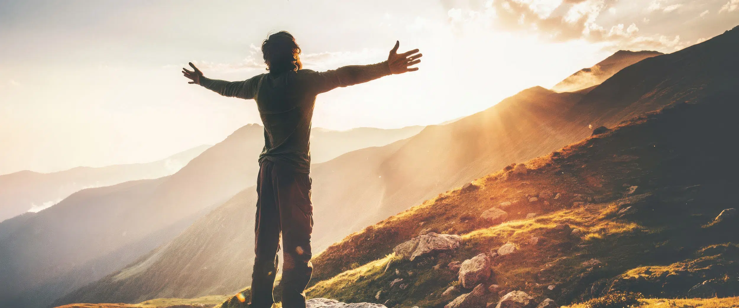 Person steht mit ausgebreiteten Armen auf einem Berg und blickt in den Sonnenaufgang über einer Berglandschaft.