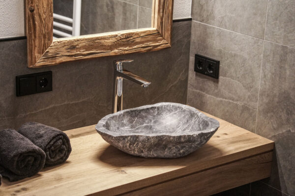 Modernes Badezimmer im Chalet-Stil mit rustikalem Holzspiegel, Naturstein-Waschbecken und minimalistischer Armatur auf einer massiven Holzplatte. Zwei gerollte, dunkle Handtücher liegen dekorativ daneben.