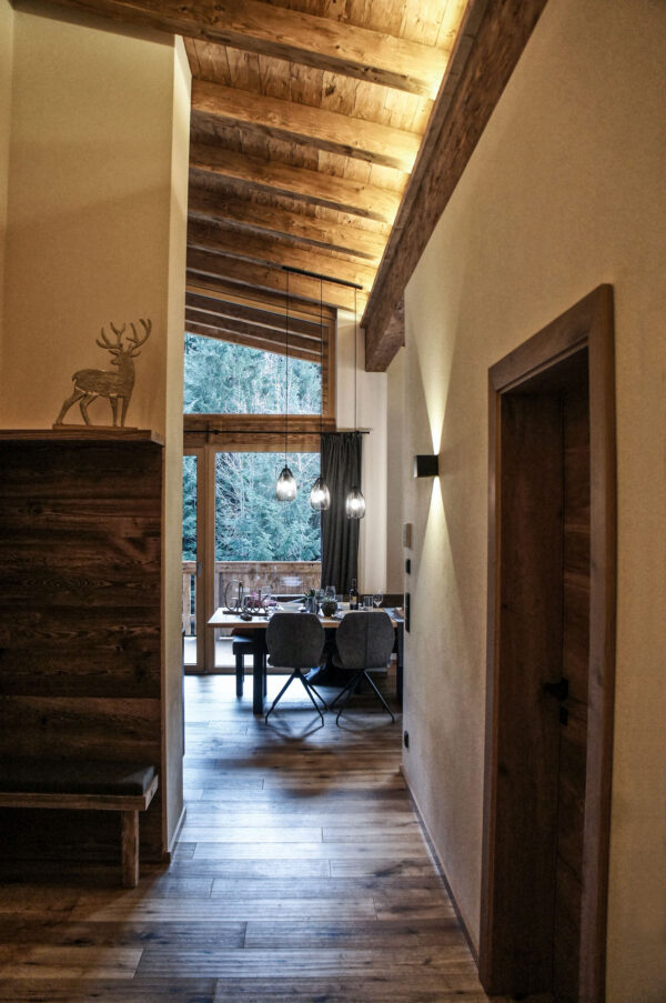 Blick vom Flur auf gedeckten Esstisch vor großer Fensterfront mit Holzdecke und Hängelampen im Chalet-Stil
