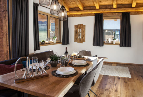 Gemütlicher Essbereich im Chalet Zeitfrei mit Holztisch, Gedeck und „Glück“-Deko im warmen Tageslicht.