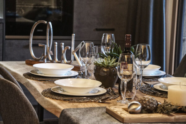 Stimmungsvoll gedeckter Esstisch aus hellem Massivholz im Chalet-Stil, arrangiert mit weißen Tellern auf geflochtenen Platzsets, Weingläsern und Besteck. In der Mitte steht eine dekorative Metallschrift „Glück“, flankiert von einem kleinen Sukkulenten-Arrangement und einer Flasche Rotwein. Ergänzt wird das Ensemble durch eine Kerze und natürliche Dekoelemente auf einem Holzbrett. Im Hintergrund moderne, dunkle Küchenfronten.