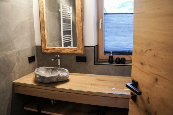 Waschtisch mit Steinbecken und Holzrahmenspiegel vor Fenster mit Sichtschutz und grauen Fliesenwänden